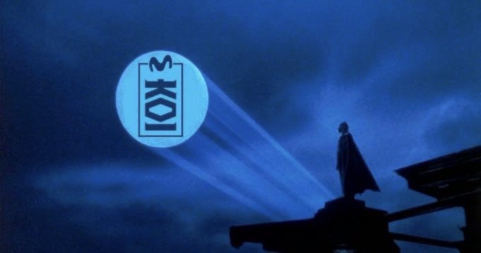 A silhouetted figure stands on a rooftop projecting a spotlight displaying the letters "SKOI" into the night sky, visually teasing an upcoming announcement about the project KOI being made from Japan.