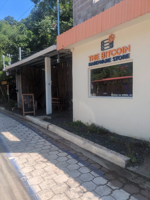 A storefront in El Salvador displays a prominent sign reading "THE BITCOIN HARDWARE STORE" in orange lettering, alongside a chalkboard advertisement for "GOT SATOSHI?" with a "B+5%" graphic. The image illustrates El Salvador's pioneering adoption of Bitcoin as legal tender, with a dedicated retail location for Bitcoin-related merchandise.