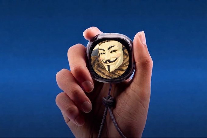 A hand holds up a smartwatch displaying the Anonymous (Guy Fawkes) mask logo against a blue background, with a braided lanyard attached.