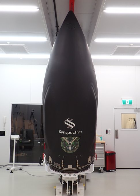 A black rocket nose cone featuring the 'Synspective' logo and emblem stands vertically in a clean laboratory or assembly facility.
