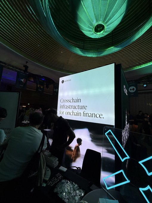 A trade show or conference display features a large illuminated sign reading 'across Crosschain infrastructure for onchain finance,' set under a green-lit domed ceiling.
