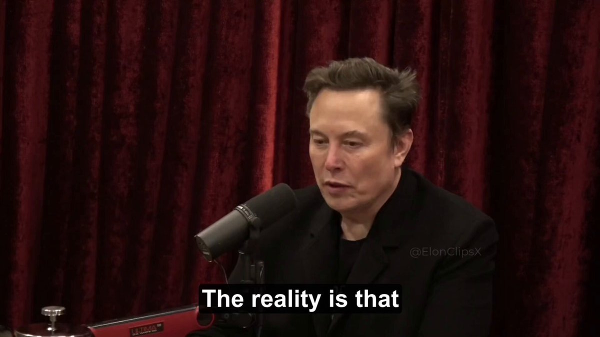 The image features an interview with a man, presumably Elon Musk, who is speaking into a microphone. He is wearing a black suit and appears to be deep in thought or contemplation. The background of the image shows a red curtain, which contrasts with the gray wall behind him. There are also some text elements present in the image that read "The reality is that our elected officials have very little power relative to the bureaucracy until DOGE is a threat to the bureaucracy". This statement seems to be related to the interview or the context of the conversation.