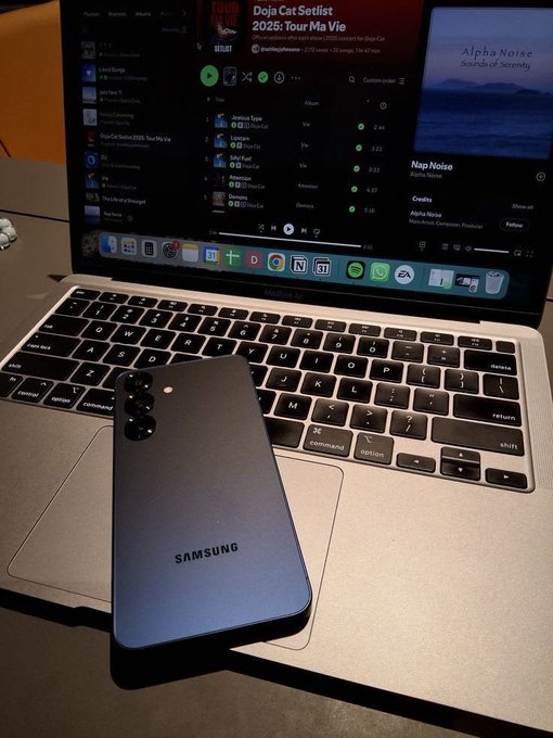 A silver Samsung smartphone resting on the keyboard of an open MacBook laptop, which shows the Spotify app playing the album "Doja Cat Setlist 2025: Tour Ma Vie."