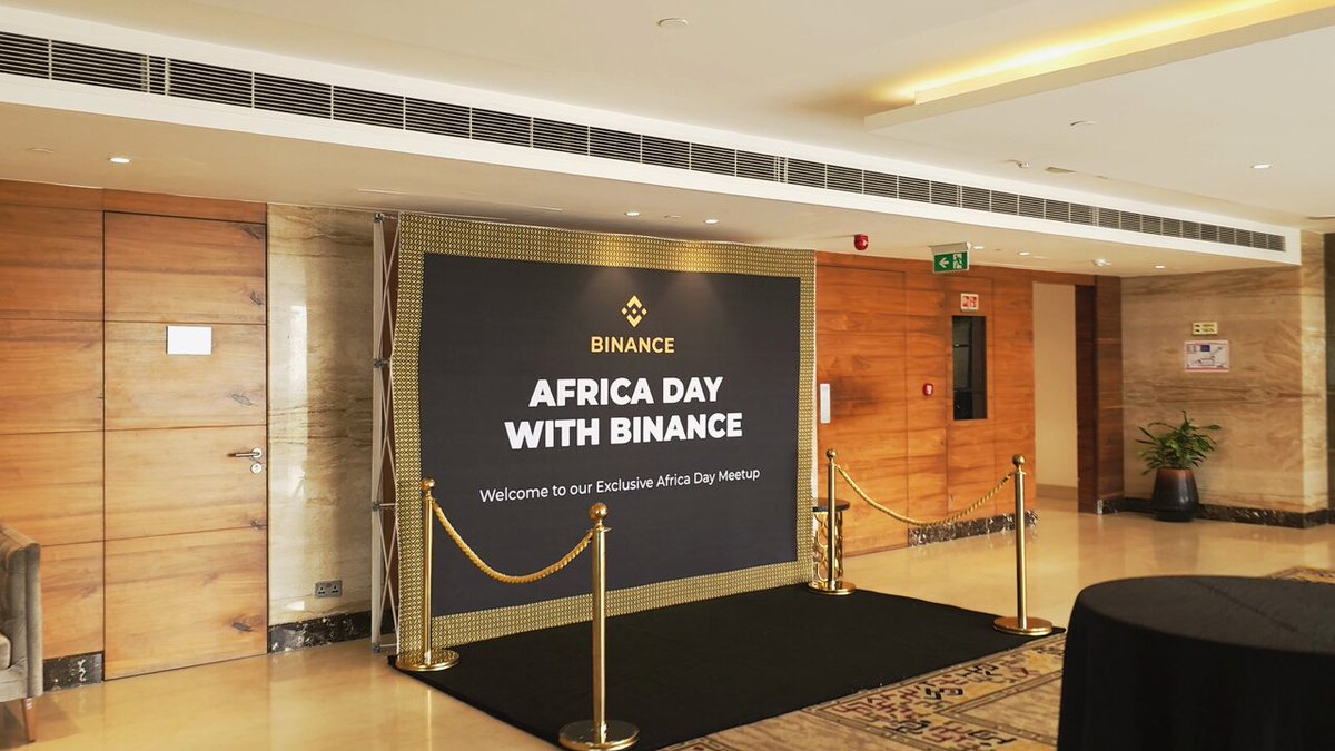 The image shows a large black banner with gold lettering that reads "Africa Day With Binance", indicating an event related to Africa or the Binance cryptocurrency platform. The banner is displayed prominently, possibly at a convention center or similar venue. In addition to the banner, there are two potted plants in the scene - one on the left side and another on the right side of the image. A dining table can also be seen towards the bottom part of the photo.