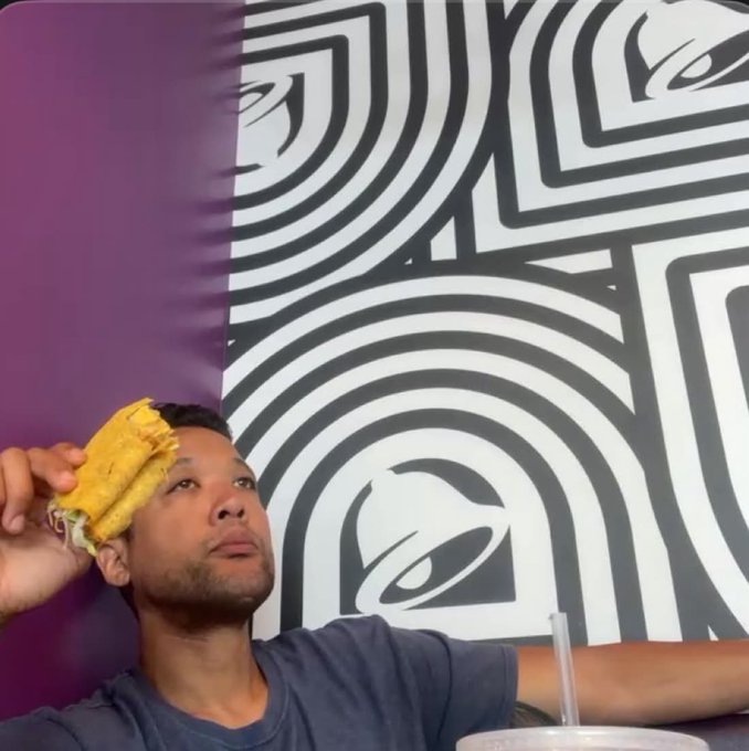 The image features a man with black hair wearing a blue shirt, holding a taco over his head as if he's about to eat it or make a funny face for the camera. The caption of the image reads "Who will be the one to step up to the Doginal Dog's Bear Market Games? I completed 20 tacos out of 36 from Taco Bell for the wizzy community Looking forward to the next competitors willing to step up to challenge". This suggests that the man is participating in a competition or event related to fast food, specifically at Taco Bell. The image captures his humorous and lighthearted approach to the situation, possibly trying to make others laugh with his silly actions.