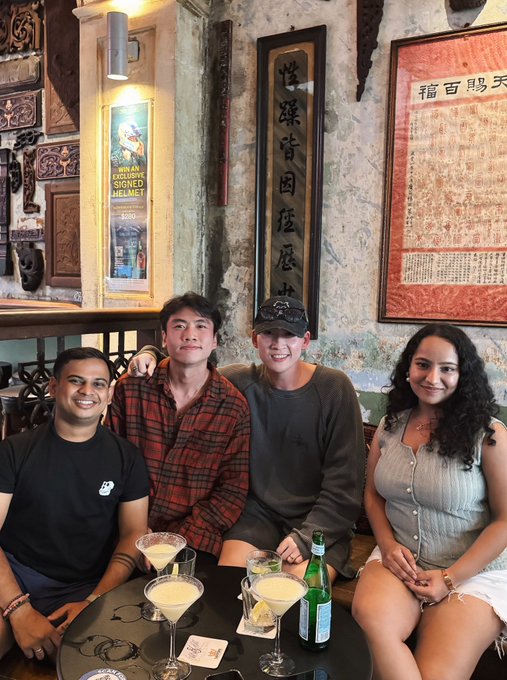 Four friends smiling for a photo at a bar, seated around a table with cocktail glasses, beneath framed Chinese calligraphy and a poster reading 'HAINAN HEROINE SIGNED HELMET 400m'.