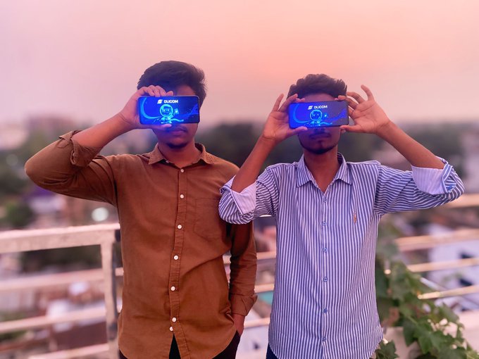 Two young men hold smartphones in front of their eyes on a rooftop at sunset, with the "DLICOM" logo visible on their screens as they pose playfully together.