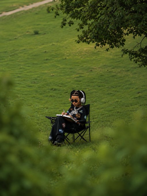 A person wearing headphones and sunglasses sits in a folding chair on a lush green field, sketching in a notebook. The scene captures a peaceful, creative "gm, happy Thursday" moment of solitary reflection in nature.