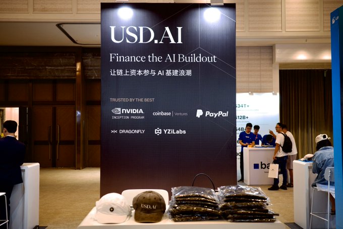 USD.AI is exhibiting at MoneyFrontier Hong Kong, with their booth banner reading "USD.AI - Finance the AI Buildout / 让链上资本参与 AI 基建浪潮" and listing partners like NVIDIA Inception, Coinbase Ventures, PayPal, Dragonfly, and YZi Labs, alongside branded merchandise displayed on the table. The accompanying post highlights their presence at the event, inviting attendees to visit and connect with the team.