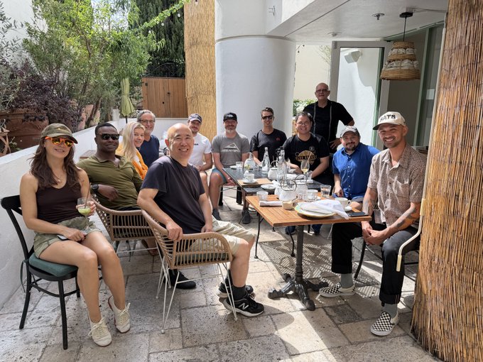 A diverse group of about eleven people gather around an outdoor table on a sunny patio, enjoying drinks and conversation together, with one person wearing a "BTS" cap. The relaxed, convivial gathering of friends enjoying a meal outdoors perfectly complements the post's sentiment about the simple joy of spending quality time with Bitcoin friends.