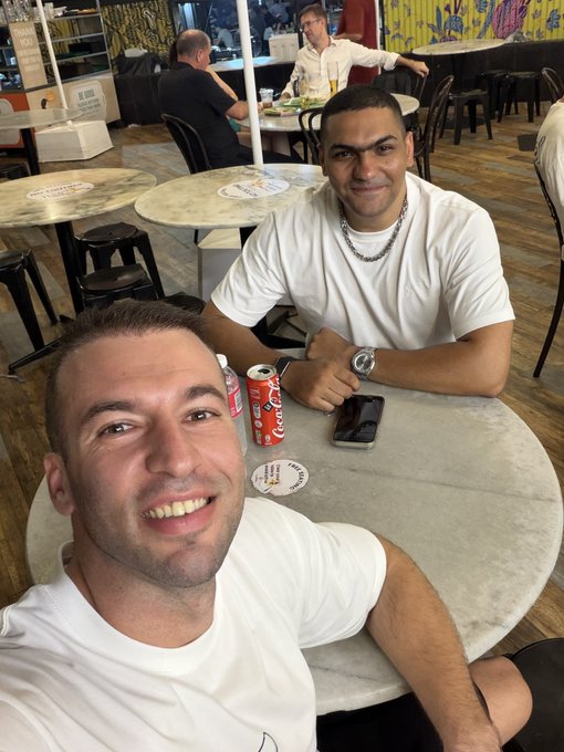 Two men sit at a round table in a restaurant, both wearing white t-shirts, with two red soda cans, a smartphone, and a lanyard visible on the table.