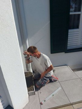 A man in a white t-shirt kneeling on a patio and working on a wall outlet with tools on the ground.