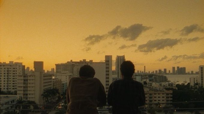 Silhouettes of two people sitting together on a rooftop, looking out over a city skyline at sunset with orange and yellow skies.