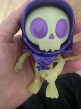 A small plastic figurine of a skeleton with a purple skull-like face and hood is held in someone's hand.