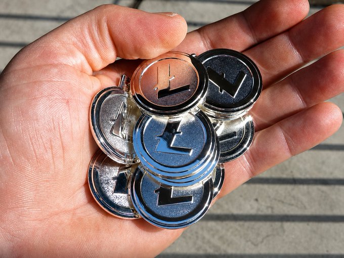 A hand cradles a stack of silver Litecoin ($LTC) physical coins, with the prominent "L" emblem visible on each coin, visually representing the post's metaphor of Litecoin as the "spending account" counterpart to Bitcoin's role as long-term wealth storage.