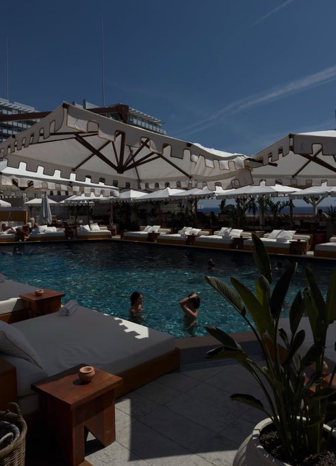 The image shows a large swimming pool with white umbrellas providing shade for people enjoying the water. There are two individuals in the pool, one of them wearing a bathing suit. The pool area also features several potted plants placed around the space, adding greenery and enhancing the ambiance. In the background, there is a building visible behind the pool, suggesting that this location might be near an urban setting or a resort with various amenities for guests to enjoy.
