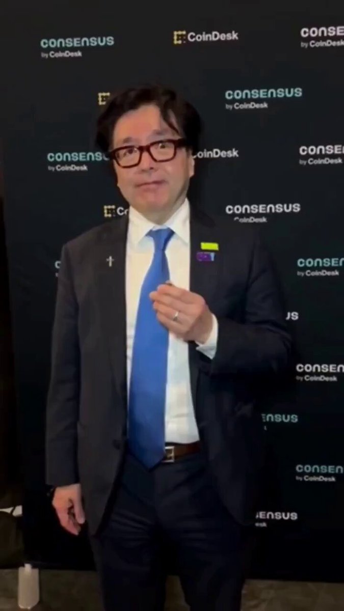The image features a man dressed in a suit and tie standing against a black background with white text. The man has glasses and is wearing a blue tie. He appears to be giving a speech or presentation, as indicated by his body language and facial expressions. In the background, there are several logos, including one for "Consensus" and another for "CoinDesk". These logos suggest that this event might be related to cryptocurrency or financial news. The man is also holding up a small object in front of him, which could be a microphone or some other equipment used during his presentation.