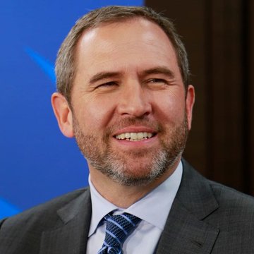 Headshot of a smiling middle-aged man with short gray hair and a beard, wearing a dark suit, white shirt, and blue tie, photographed against a blue background.