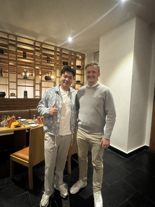 Two men pose for a photo in what appears to be a restaurant setting with wooden shelving and decorative items in the background.
