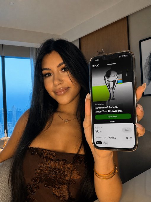 The image features a woman holding up her phone displaying a World Cup app. The app showcases a photo of the soccer trophy, indicating that she is watching or discussing the event. She also has a cupcake picture next to the trophy, suggesting that there might be a celebration related to the World Cup. The post includes information about the World Cup and possibly some details about the app's features, such as its ability to track match outcomes, check in daily, and provide insights into the cryptocurrency market.