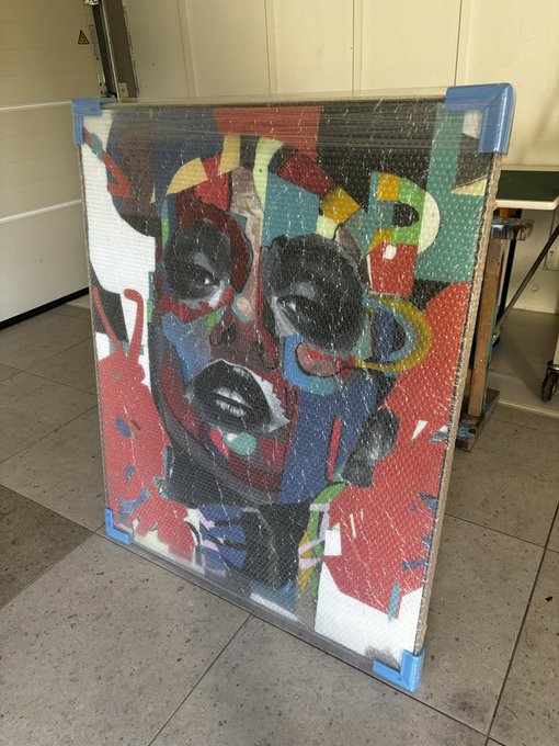 The image features a large abstract painting of a face that has been wrapped up in plastic for protection during transportation or storage. The painting is displayed prominently against a white wall, drawing attention to its vibrant colors and intricate details. The artist's signature can be seen on the bottom right corner of the frame.