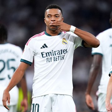 Soccer player Kylian Mbappé in a white Real Madrid jersey (with 'Emirates FLY BETTER' sponsor) making a goal celebration gesture by blowing on his fingers.