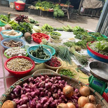 A vibrant display of fresh produce including red onions, shallots, tomatoes, garlic, chili peppers, and leafy greens is arranged in colorful baskets at an outdoor market. The caption humorously illustrates the post's narrative of a crypto trader forced to sell vegetables to recover from losses after being scammed on rewards and losing money on MoonPay transactions.