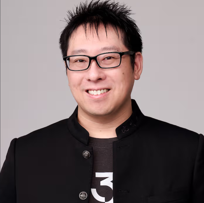 A professional headshot of an Asian man with spiky black hair and glasses, smiling while wearing a black jacket over a t-shirt.