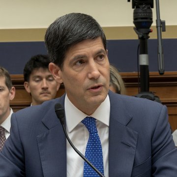 A man with graying hair in a blue suit and patterned tie speaking into a microphone, likely during a congressional or committee hearing.