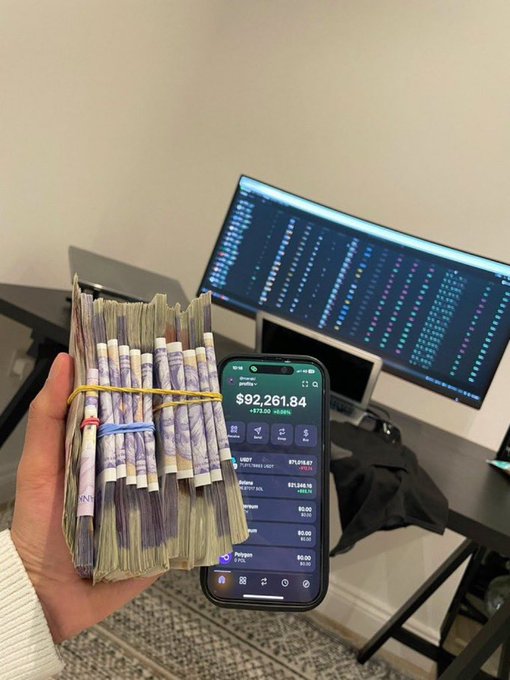 A person holds a thick stack of foreign currency bills secured with rubber bands in one hand and a smartphone displaying a Robinhood portfolio totaling $92,261.84 in the other, with a monitor showing stock charts in the background.