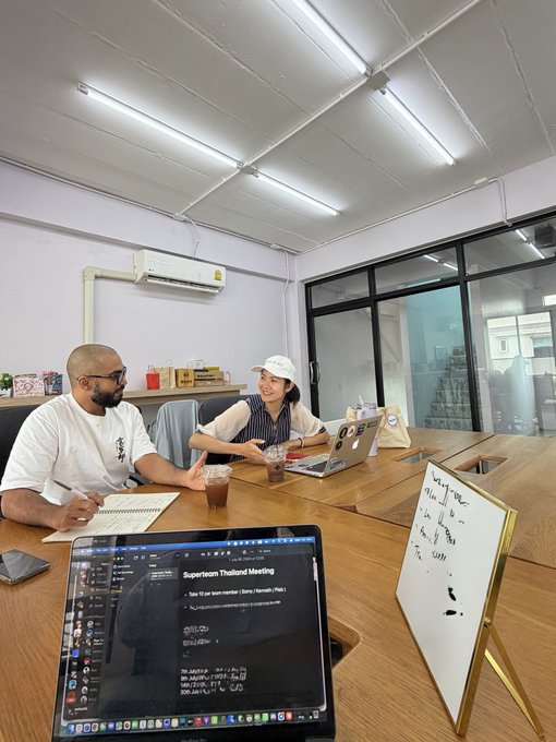 Two people having a meeting at a conference table with a laptop displaying a 'Superteam Thailand Meeting' agenda and an AirPlay screen showing code on a MacBook in the foreground.