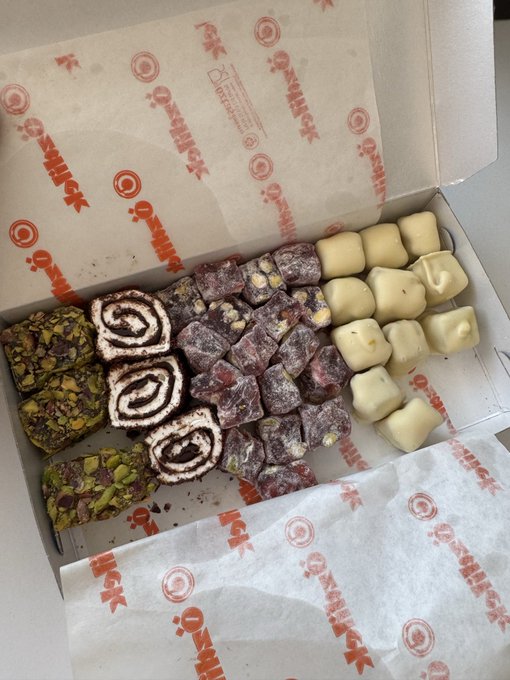 An open box of Turkish delight (lokum) containing various pieces including pistachio-topped cubes, chocolate-swirled rolls, and plain white squares, lined with branded parchment paper.
