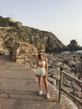 A woman in a white top and shorts standing on a coastal pathway with rocky cliffs, blue sea, and buildings visible in the background.