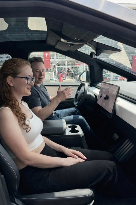 Elon Musk and a woman with glasses sit inside a vehicle, likely a Tesla Cybertruck, looking at the dashboard display.