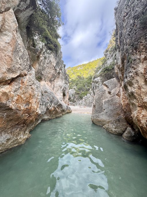 A scenic view of a turquoise river flowing through a narrow canyon surrounded by tall rocky cliffs, with green vegetation visible in the background under a partly cloudy sky.