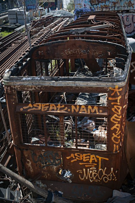 A rusty, abandoned tram or cable car sits in a derelict yard, covered in graffiti including the names 'ASFAL NAM' and 'WBO', with its roof removed and interior filled with debris.