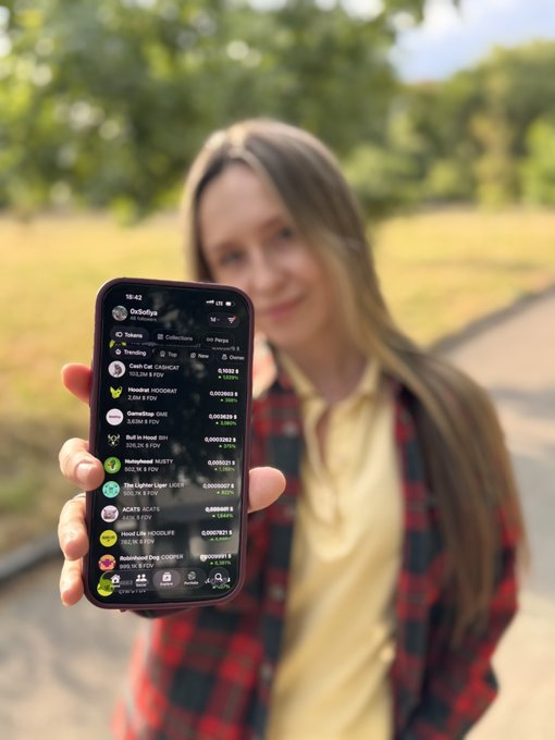 A person holding up a smartphone displaying a list-based app interface, showing trending items with various numbers and names like 'Roast Beef HOODRAY' and 'SnowTime IP68'.