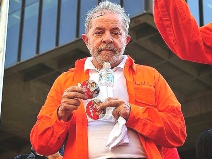 An older man wearing an orange shirt with "LULA" embroidered on it holds a water bottle and political stickers featuring Lula's face. The image humorously relates to the post's claim that Brazil hasn't won a World Cup since Lula became president, visually blaming his leadership for the national team's losses.