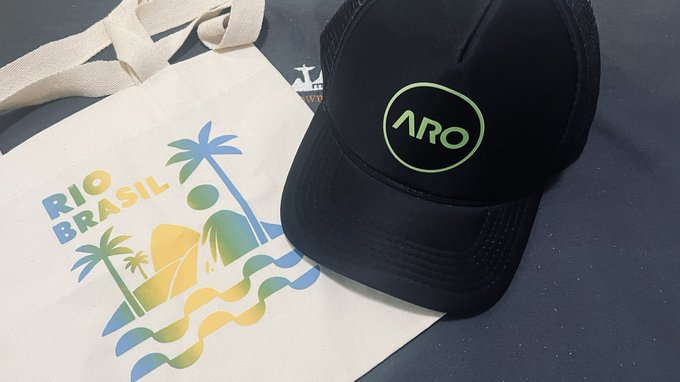 A black ARO trucker hat rests atop a canvas tote bag featuring a colorful "RIO BRASIL" tropical design with palm trees and waves, representing the brand's presence at Blockchain Rio. The promotional items align with the post's announcement of their Brazilian ambassador arriving at the event to network with Web3 projects.