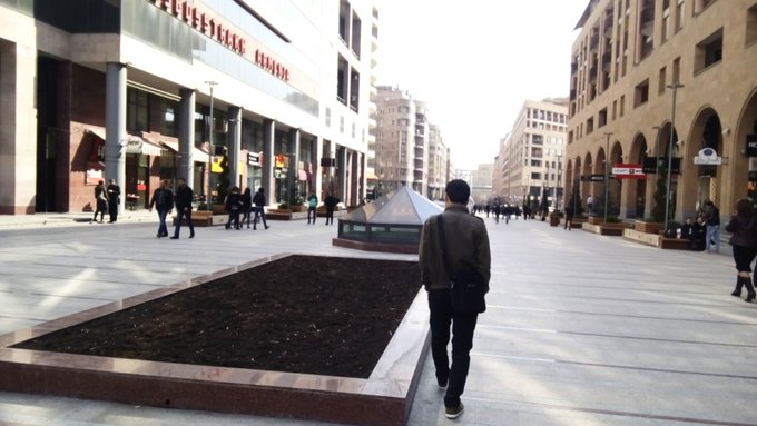 A person walks through an outdoor pedestrian plaza lined with shops and arched buildings, with a rectangular planter in the foreground.