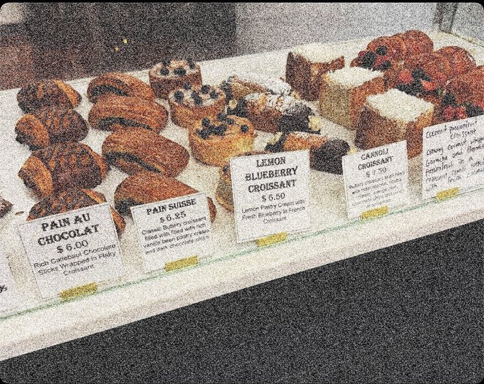 A bakery display case shows various pastries with handwritten labels including 'Pain au Chocolat $6.00', 'Pain Suisse $6.25', 'Lemon Blueberry Croissant $6.50', and 'Carrot Croissant $7.50'.