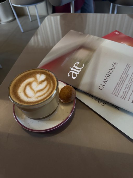 A frothy cappuccino featuring a latte art heart sits beside a small cookie on a saucer, with a folded magazine showing "ate" and "GLASSHOUSE" headlines resting on the table. The cozy café scene pairs perfectly with a relaxed "GM, happy sunday" greeting—ideal inspiration for a leisurely weekend morning.