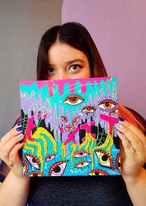 A young woman with dark hair holds a colorful, abstract piece of artwork featuring multiple stylized eyes in yellow, pink, and blue tones directly in front of her lower face against a split pink and purple background.