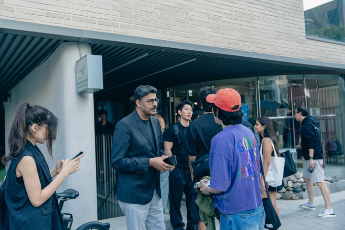 A group of casually dressed individuals stands outside a building entrance with a tiled facade; a woman in a black vest looks at her phone on the left, while a man in a black suit stands in the center, and another man wears a red cap.