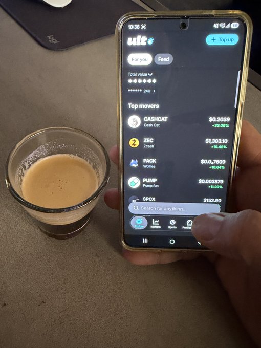 A hand holds a smartphone displaying the Ult Finance app showing 'Top movers' including CASHCAT (+33.06%), ZEC (+16.48%), PACK (+16.64%), PUMP (+13.20%), and SPCX ($152.90), next to a small glass of tea.