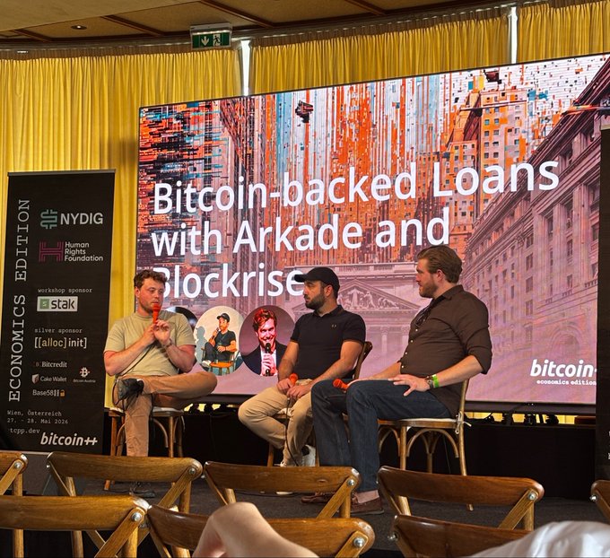 The image shows three men sitting around a table, engaged in conversation. They are positioned behind a screen displaying an advertisement for Bitcoin-backed loans with Arkade and Blockrisse. The backdrop features a cityscape, adding context to their discussion about digital currency.