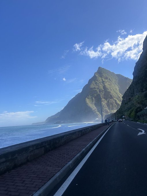 A scenic coastal highway hugs a dramatic cliffside beside the ocean under a bright blue sky with wispy clouds. The "GM" in the post likely ties to "Good Morning," with the image capturing a fresh, sunlit start to the day perfect for a morning greeting.