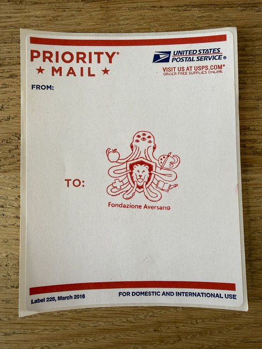 A blank USPS Priority Mail envelope addressed from "Fondazione Aversano" sits on a wooden surface, ready to be filled out and sent. The accompanying post suggests the sender is preparing to mail something to invite digital blockchain artists to a 2-week residency in Australia.