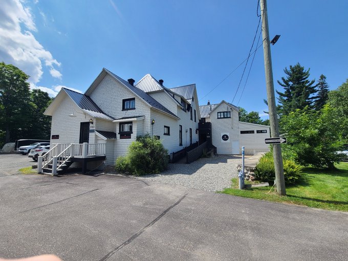 A bright, sunny daytime photo of a large white multi-story house with a metal roof, gravel driveway, and attached garage, surrounded by green trees.