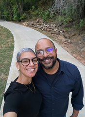 A selfie of a smiling man and woman with glasses standing on a curving paved road surrounded by trees.